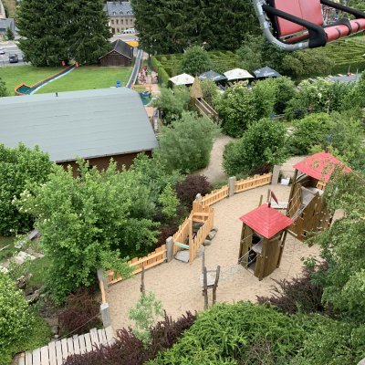 Ein Spielplatz liegt in der Natur mit einem grünen Umfeld. Kinder spielen auf einer Rutsche und erkunden die Holzhütten.