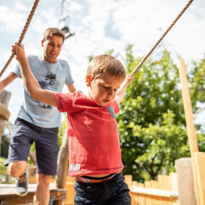 Ein Junge und sein Vater spielen im Park. Der Junge zieht an einem Seil und lächelt. Es ist ein sonniger Tag.