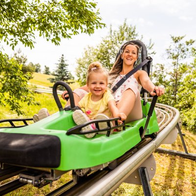 Eine Mutter fährt mit ihrem kleinen Kind auf einer Sommerrodelbahn in der Natur an einem sonnigen Tag.