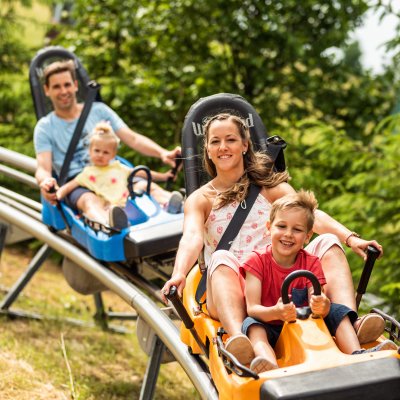 Eine Familie fährt auf einer Sommerrodelbahn in den Bergen. Es sieht nach viel Spaß aus. Kinder sitzen vorne und lächeln.