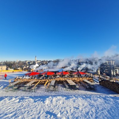 Ein Skigebiet zeigt Bereiche mit Schnee, Tischen, Stühlen und Dampfsäulen. Menschen nutzen die Winterlandschaft.