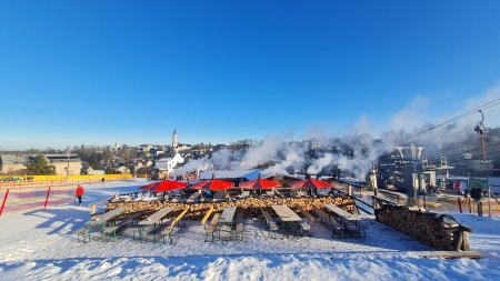 Ein Skigebiet zeigt Bereiche mit Schnee, Tischen, Stühlen und Dampfsäulen. Menschen nutzen die Winterlandschaft.