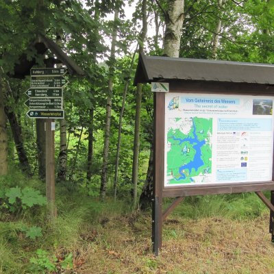 Informationstafel und Wegweiser stehen an einem Waldweg. Eine Karte zeigt Wanderrouten zum Wasserlehrpfad Eibenstock