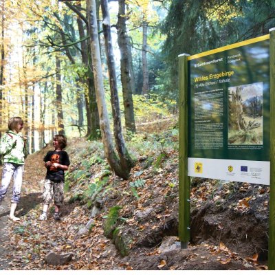 Zwei Kinder stehen neben einem Schild im Wald der Erzgebirge und schauen neugierig darauf. Es ist Herbst.