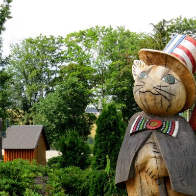 Eine Holzskulptur einer Katze steht in einem Park. Um die Statue sind Bäume und Sträucher. Im Hintergrund ist ein kleiner Holzschuppen zu sehen.