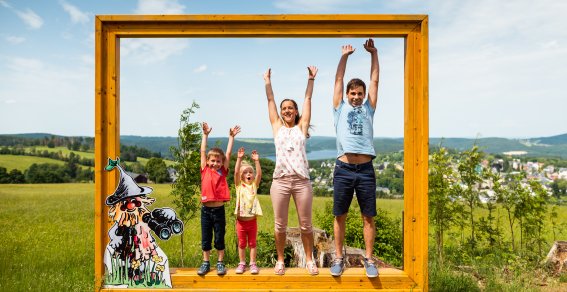 Familie Bilderrahmen Wurzelrudis Erlebniswelt Eine Familie steht glücklich in einem großen Holzrahmen auf einem Feld. Die Kinder und die Mutter lächeln.