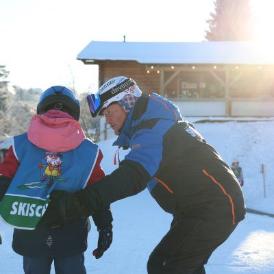 Ein Skilehrer hilft einem Kind beim Skifahren in einem Wintersportgebiet. Die Sonne scheint, und Schnee liegt auf dem Boden.