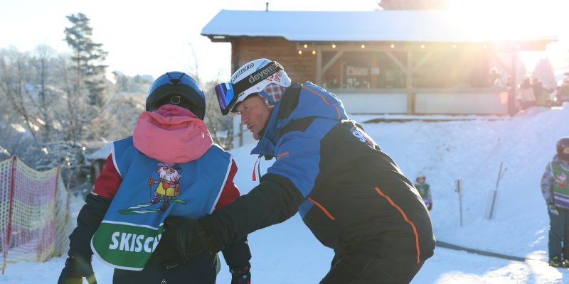 Ein Skilehrer hilft einem Kind beim Skifahren in einem Wintersportgebiet. Die Sonne scheint, und Schnee liegt auf dem Boden.