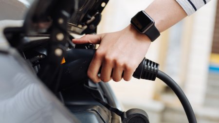 Eine Hand steckt ein Ladekabel in ein Elektroauto. Es ist Tag und das Auto steht an einem Ladepunkt.