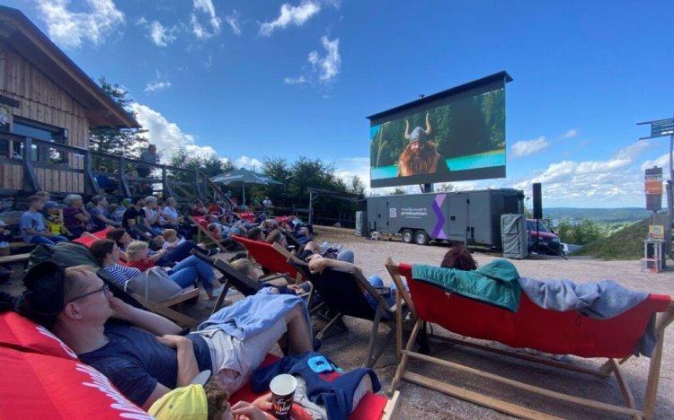Menschen sitzen auf Liegestühlen und sehen einen Film im Freien. Das Wetter ist sonnig und die Stimmung ist entspannt. Moviequidi-Eibenstock