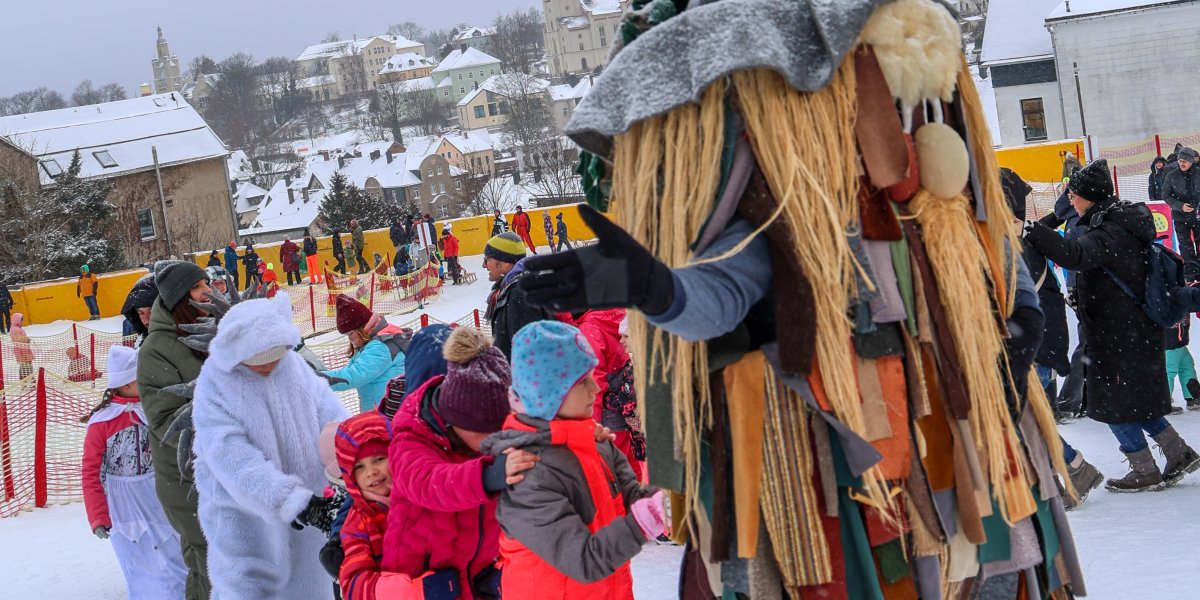 Kinder stehen im Schnee und spielen mit einem großen Maskottchen. Im Hintergrund sind Gebäude und eine Kirche sichtbar.