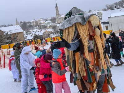 Kinder stehen im Schnee und spielen mit einem großen Maskottchen. Im Hintergrund sind Gebäude und eine Kirche sichtbar.