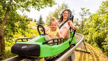 Zwei Kinder fahren mit einer Schlittenbahn auf einer Strecke im Freien an einem sonnigen Tag zwischen Bäumen.