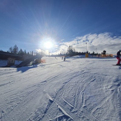 Menschen fahren Ski und Snowboard auf einer verschneiten Piste unter einem blauen Himmel. Die Sonne scheint hell.